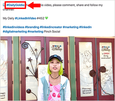 Toto je snímka obrazovky, ktorá ilustruje, ako Goldie Chan používa hashtagy v texte svojich videonahrávok na LinkedIn. Červené popisy poukazujú na hashtag #DailyGoldie v texte, ktorý je jedinečný pre jej príspevky k videu a pomáha jej sledovať zdieľania. Tento príspevok obsahuje aj ďalšie príslušné hashtagy, ktoré pomáhajú ľuďom nájsť jej video, vrátane #LinkedInVideo. Na videozázname stojí Goldie pred niektorými dverami na displeji World of Disney. Je to ázijská žena so zelenými vlasmi. Oblečenú má čiernu šiltovku LinkedIn, čierny náhrdelník s obojkom, ružovú košeľu s potlačou macaron a modro-biele sako.